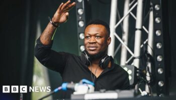 Benga looks out from behind a DJ desk on stage, his had raised