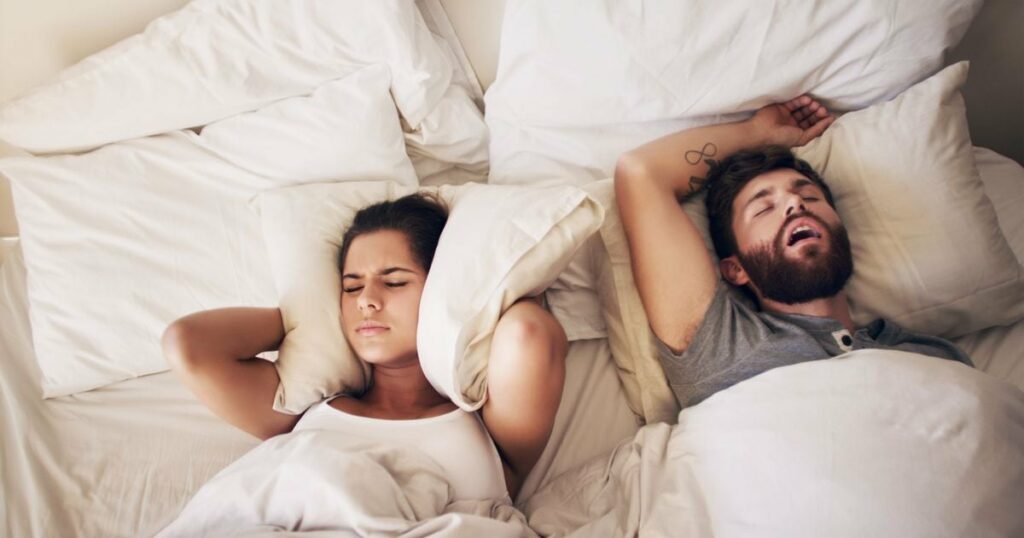 Une jeune femme se bouche les oreilles avec un coussin pendant que son mari ronfle dans le lit.