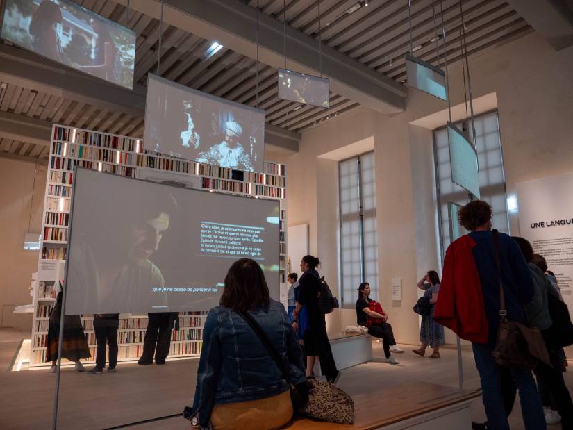 photo 5 : Plusieurs visiteurs observent différentes ​installations ⁢dans une salle d’exposition. Au centre,​ un grand écran projette une vidéo. ⁣Des écrans ⁣plus petits sont suspendus au plafond, ‌diffusant⁣ également des vidéos.