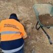 Photo 1 : Un homme porte un gilet de sécurité orange, il est agenouillé sur un sol terreux, et effectue des fouilles archéologiques. À côté de lui, une brouette contenant de la terre est posée sur le sol.