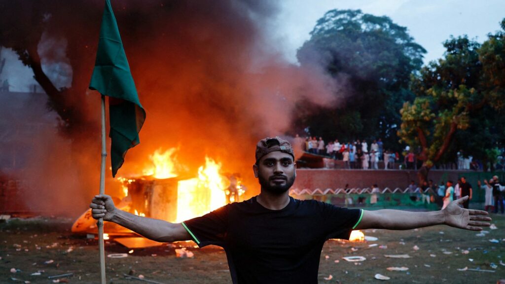 A man holding a Bangladesh flag stands in front of a vehicle that was set on fire at the Ganabhaban, the Prime Minister's residence, after the resignation of PM Sheikh Hasina in Dhaka, Bangladesh, August 5, 2024. REUTERS/Mohammad Ponir Hossain TPX IMAGES OF THE DAY