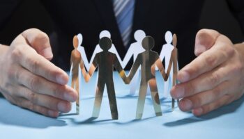 Businessman's Hands Protecting Team Of Paper People On Desk