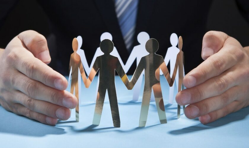 Businessman's Hands Protecting Team Of Paper People On Desk