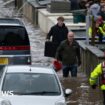 People wade through water on a street in Pontypridd, Wales, with an emergency worker in the foreground and two vehicles also partially submerged.