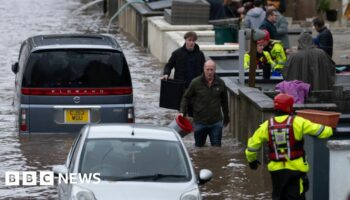 People wade through water on a street in Pontypridd, Wales, with an emergency worker in the foreground and two vehicles also partially submerged.