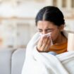 Cold And Flu Symptoms . Sick young indian woman covered in blanket blowing her running nose in a tissue while sitting on couch in living room, ill lady drinking hot tea, feeling unwell at home